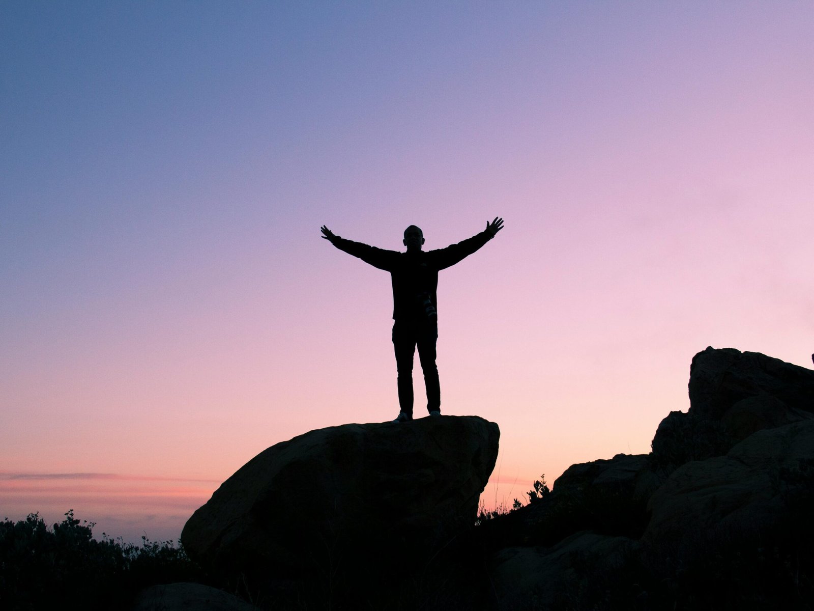 Silhouette of a man on a rock with arms raised, embracing the sunset sky.